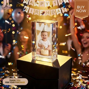 May include: A clear crystal photo block displaying a baby with a birthday cake and a party hat. The block is on a black box with a gold top. The words "HAPPY 1ST BIRTHDAY" are in the background. The image also includes the text "BUY NOW".