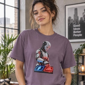 May include: A dusty purple short-sleeve t-shirt featuring a graphic of a white and silver robot pressing a red button labeled HUMAN EXTINCTION next to a blue button with a stick figure icon. The robot has red accents on its mechanical body.
