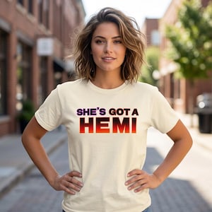 May include: A cream-colored short-sleeve t-shirt featuring the text "SHE'S GOT A HEMI" printed across the chest. The lettering "SHE'S GOT A" appears in a dark purple, while the word "HEMI" is displayed in a bold, gradient orange and red font.