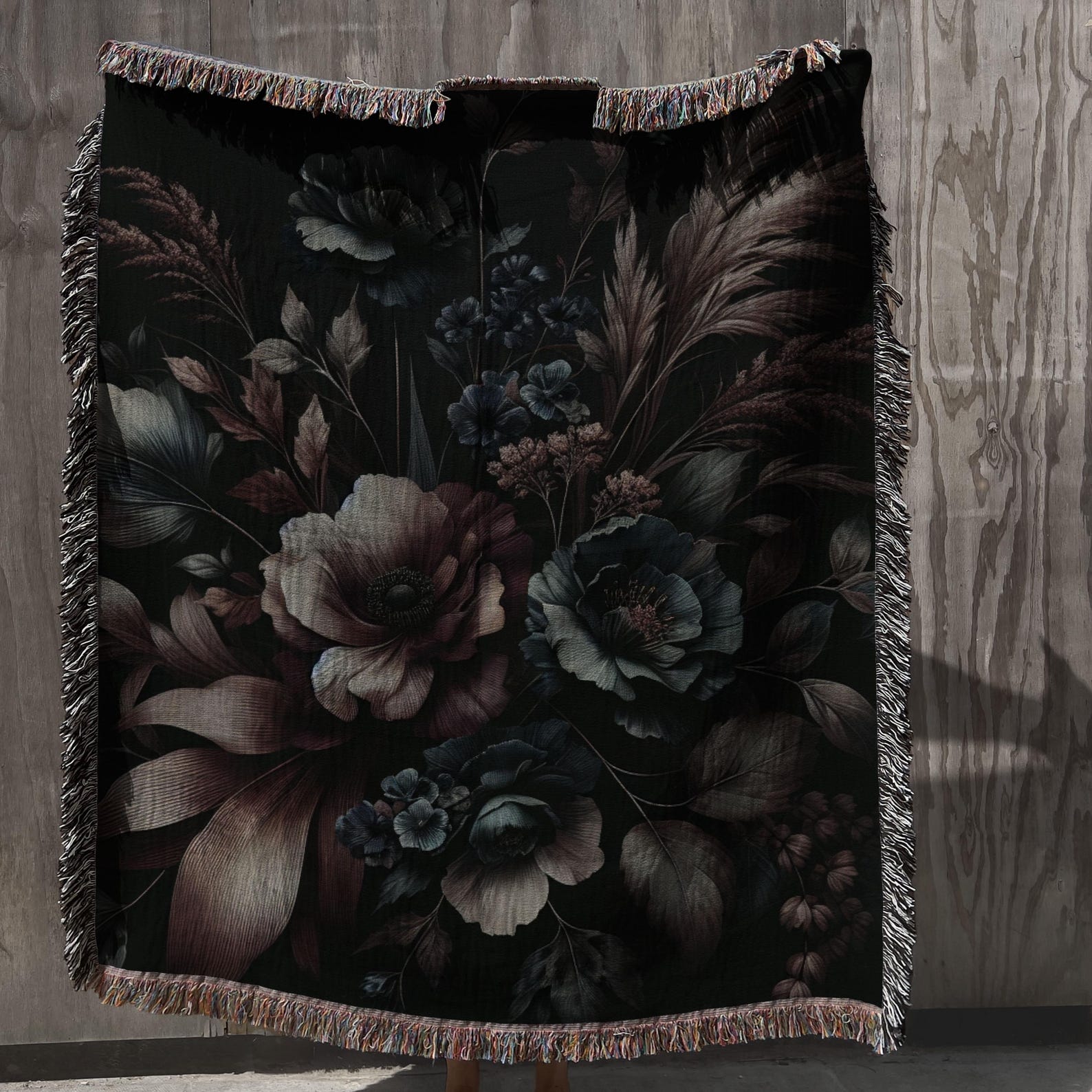 Black woven throw blanket with baroque-style floral design in muted tones and fringe trim, hanging against wood panel backdrop