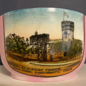 May include: A ceramic bowl featuring a colorful illustration of Elsinore Tower in Eden Park, Cincinnati, Ohio. The scene depicts a stone castle-like structure with a tall tower and a flag, set against a soft sky. The bowl has a light pink rim.
