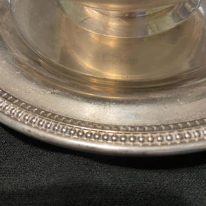 May include: A close-up of a silver-colored metal serving dish. The dish has a decorative beaded edge and a tiered design. The surface of the dish reflects light, showing a shiny, metallic appearance. The background is a dark, textured surface.