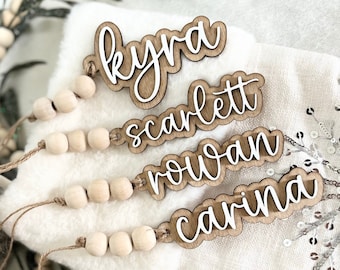 May include: Four wooden name tags with white script lettering: Kyra, Scarlett, Rowan, and Carina. Each tag is attached to a string of wooden beads. The tags are layered on a white, fluffy surface, with decorative greenery in the background.