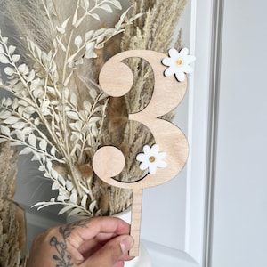 May include: A wooden number three cake topper with two white daisy accents. The number is made of light-colored wood and has a long, thin stem for insertion into a cake. The daisies have a yellow center.