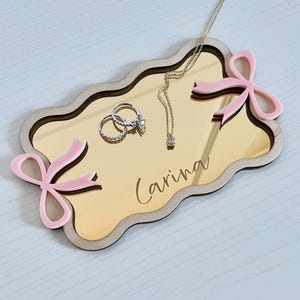 May include: A decorative jewelry tray with a gold mirrored surface and a light wood border. The tray features two pink bow accents and the name "Carina" in script. Two rings and a necklace with a pendant are displayed on the tray.