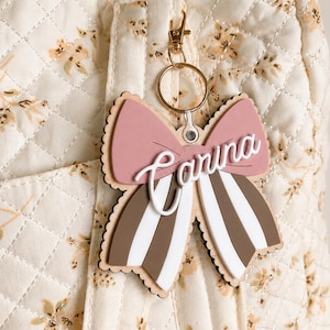 May include: A decorative bow-shaped keychain featuring a dusty rose top layer, brown and white striped bottom layers, and the name Carina in white script lettering. The accessory includes a gold-toned metal lobster clasp and a circular ring.