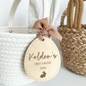 May include: A white woven basket with a handle and bunny ears, next to a wooden Easter egg tag. The tag is engraved with the name "Kolden's", "FIRST EASTER 2025", and a bunny illustration. A brown ribbon is tied to the tag.