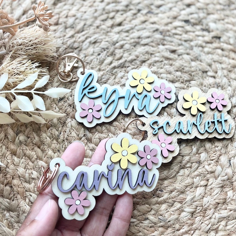 May include: Three wooden keyrings with floral accents. The keyrings are painted in pastel colours and feature the names "Kyra", "Scarlett", and "Carina".