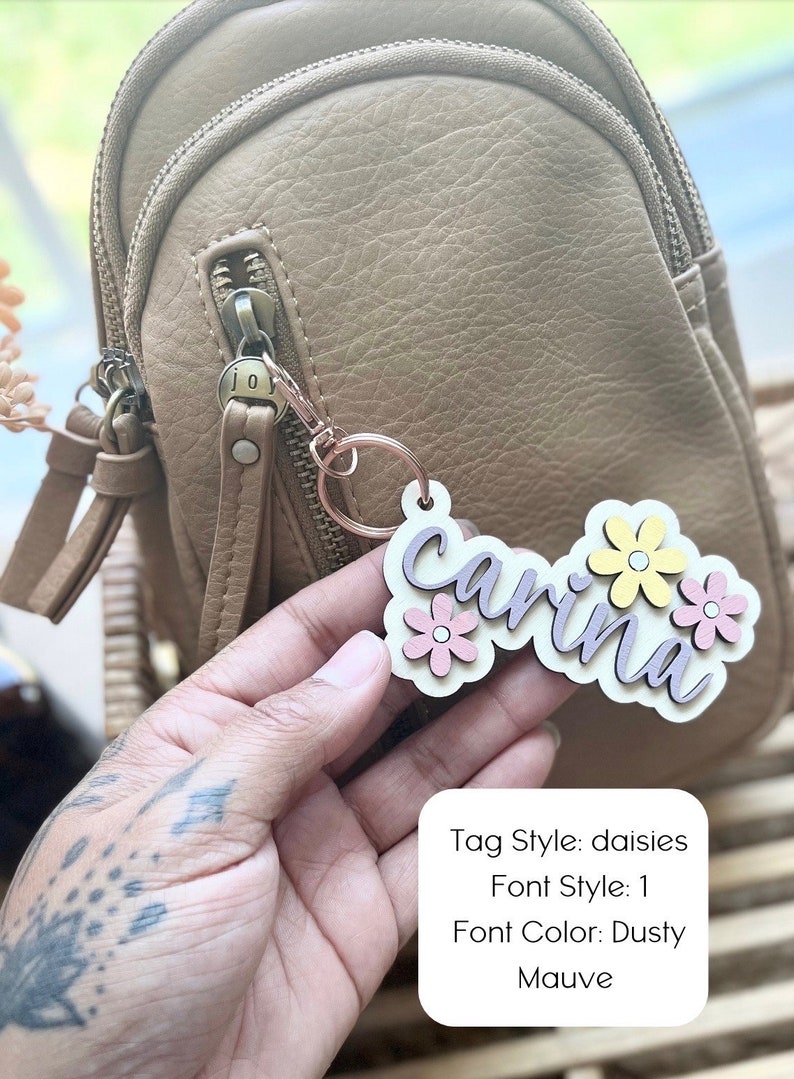 May include: A wooden keyring with the name "carina" written in a dusty mauve font. The keyring is decorated with three pink and yellow daisies. The keyring is attached to a brown leather bag.