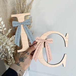May include: Two wooden letters, "J" and "C", are displayed. The "J" has a light blue ribbon with a floral pattern, while the "C" has a pink ribbon with a similar floral design. The letters are held against a neutral background.