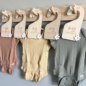 Puede incluir: Un organizador de ropa de bebé de madera con cinco perchas etiquetadas "NB newborn", "0-3 meses", "3-6 meses", "6-9 meses" y "9-12 meses". Cada percha tiene un acento de flor blanca y sostiene un body en varios colores: melocotón, amarillo y verde salvia.
