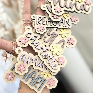May include: A set of wooden keyrings with different names, each decorated with pink and yellow flowers. The names on the keyrings are "Nina", "Reagan", "Yula", "Charlotte", "Amu", and "Mari".