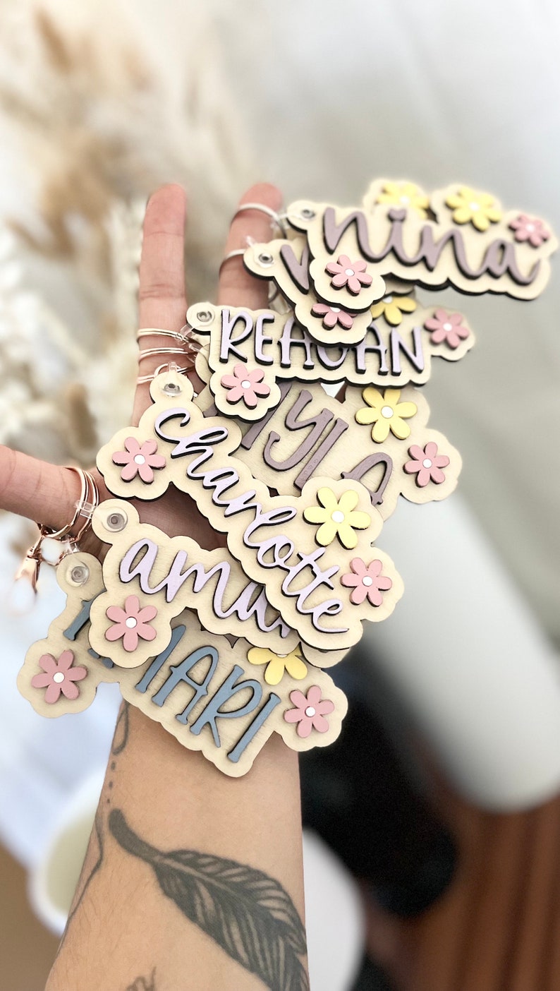 May include: A set of wooden keyrings with different names, each decorated with pink and yellow flowers. The names on the keyrings are "Nina", "Reagan", "Yula", "Charlotte", "Amu", and "Mari".