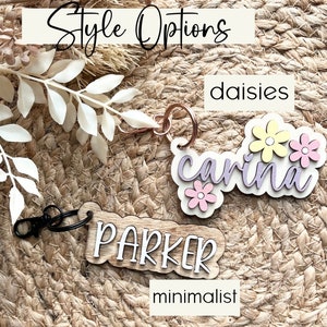 May include: Two wooden keychain tags with different styles. The first tag is a light wood with the name "carina" in purple script with three yellow and pink flowers. The second tag is a darker wood with the name "PARKER" in white script. The text "daisies" is above the first tag and "minimalist" is below the second tag.