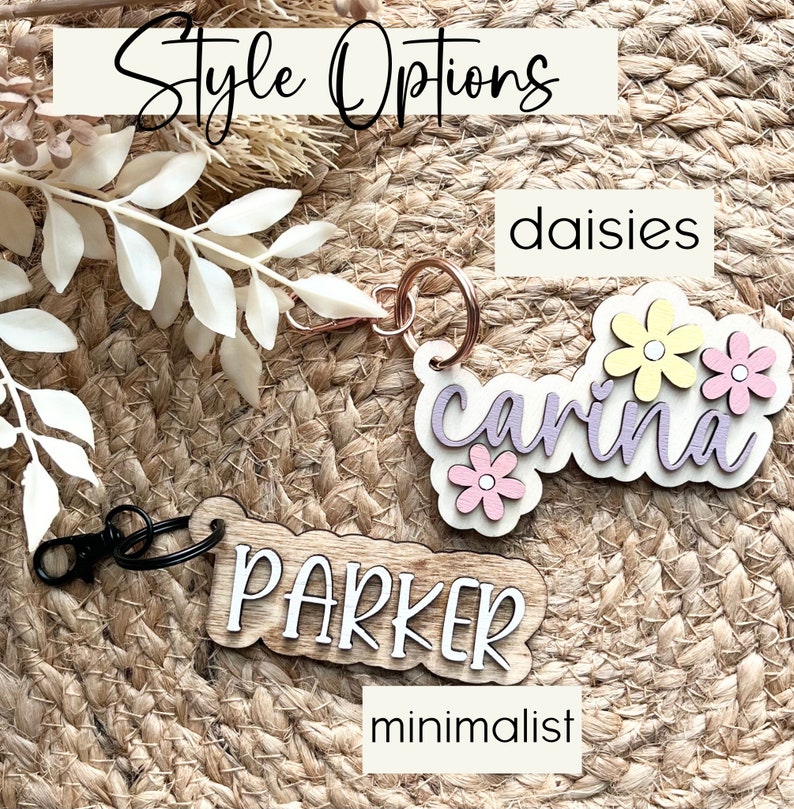 May include: Two wooden keychain tags with different styles. The first tag is a light wood with the name "carina" in purple script with three yellow and pink flowers. The second tag is a darker wood with the name "PARKER" in white script. The text "daisies" is above the first tag and "minimalist" is below the second tag.