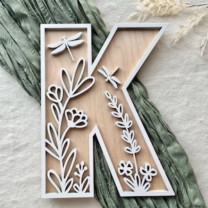 May include: Wooden letter "K" with a white border and floral and dragonfly cutouts. The letter is made of light-colored wood and features white dragonfly and floral designs. The background is a light green fabric.