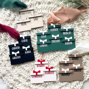May include: Wooden gift box ornaments in various colors, each with a white bow and personalized name. Names include Mom, Dad, Chris, Carina, Kyra, Kirsten, Honey, Merlin, Nathan, Ryker, Jonah, Theo, Daddy, Mommy, Rowan, Scarlett, Courtney, Goji, Ciarah, and Baby A 2026.