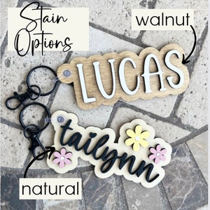 May include: Two wooden keychains with the names "Lucas" and "Tailynn" in black lettering. The keychain with "Lucas" is stained walnut and the keychain with "Tailynn" is stained natural. Both keychains have a black metal key ring.