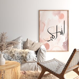 May include: A watercolor print with a pink and brown background featuring the Arabic calligraphy "Allah" in black. The print is framed and hanging on a white wall above a couch and chair.
