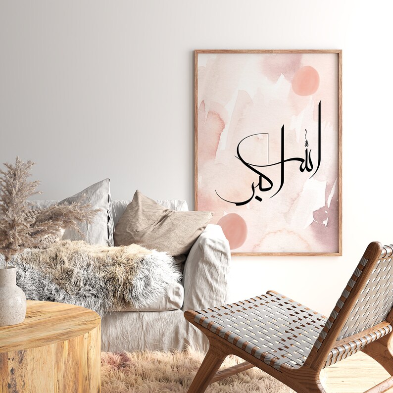 May include: A watercolor print with a pink and brown background featuring the Arabic calligraphy "Allah" in black. The print is framed and hanging on a white wall above a couch and chair.