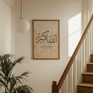 May include: A framed print with Arabic calligraphy in brown ink. The text reads "ALLAHUAKBAR" above "ALLAH IS THE GREATEST" in a serif font. The print is in a brown wooden frame, hanging on a white wall near a staircase.