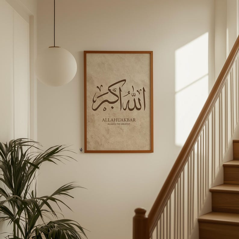 May include: A framed print with Arabic calligraphy in brown ink. The text reads "ALLAHUAKBAR" above "ALLAH IS THE GREATEST" in a serif font. The print is in a brown wooden frame, hanging on a white wall near a staircase.