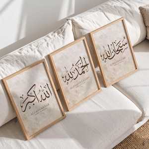 May include: Three framed art prints with Islamic calligraphy in brown script on a beige background. The wooden frames are rectangular. The prints feature the phrases: ALLAHUAKBAR, ALHAMDULILLAH, and SUBHANALLAH, with English translations below.