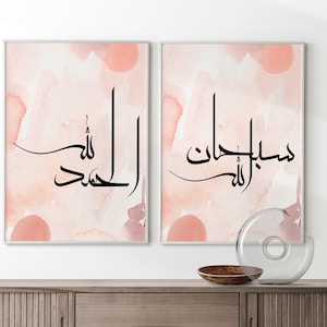 May include: Two framed prints with a watercolor background in shades of pink and white. The prints feature Arabic calligraphy in black ink. The text on the left print reads "Alhamdulillah" and the text on the right print reads "Masha'Allah".