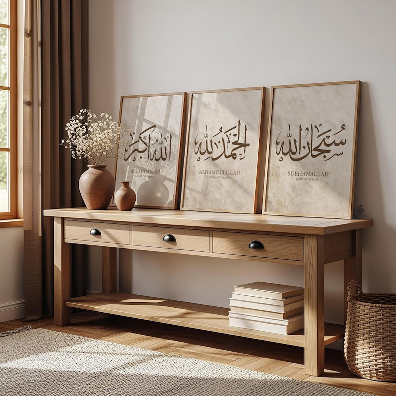 May include: Three framed Islamic calligraphy art prints with brown wooden frames are displayed on a light brown wooden console table. The prints feature Arabic script in a dark brown color. A vase with dried flowers and two smaller vases are on the table.
