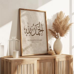 May include: A framed print with Arabic calligraphy, reading "Alhamdulillah, Praise be to Allah." The artwork is in a wooden frame, displayed on a wooden cabinet. A vase with dried pampas grass and decorative items complete the scene.