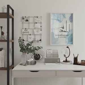 May include: A white desk with a black metal shelf unit, a globe, a wire mesh organizer, a gray notebook, a camera, a vase with green leaves, and a framed print with the Arabic text "Alhamdulillah" and the English translation "Praise be to Allah".