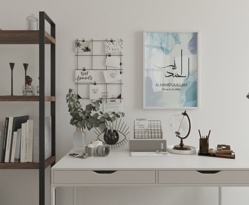 May include: A white desk with a black metal shelf unit, a globe, a wire mesh organizer, a gray notebook, a camera, a vase with green leaves, and a framed print with the Arabic text "Alhamdulillah" and the English translation "Praise be to Allah".