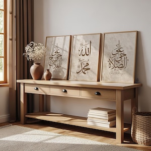 May include: Three framed prints with Arabic calligraphy in brown tones are displayed on a light wood console table. The table has three drawers with black knobs and a lower shelf. A vase with dried flowers and a woven basket are also visible.