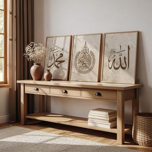 Puede incluir: Tres impresiones enmarcadas de caligrafía islámica en tonos marrones. Las impresiones se exhiben en una consola de madera clara con tres cajones y un estante inferior. Un jarrón con flores secas y dos jarrones más pequeños están sobre la mesa. También hay una cesta tejida y una pila de libros.