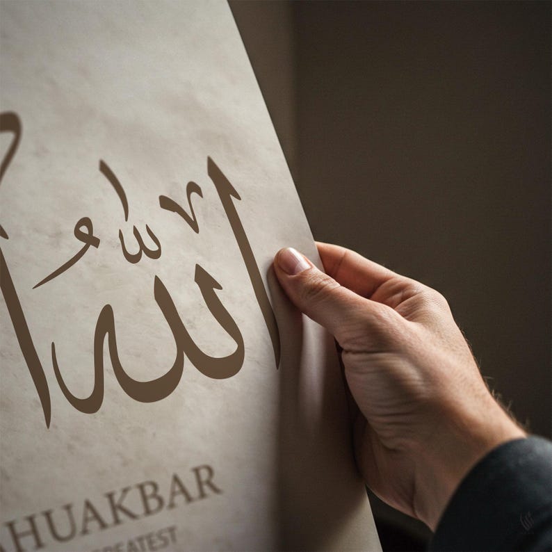 May include: A beige print featuring Arabic calligraphy in brown ink. The artwork is held by a hand, with the text "ALLAHUAKBAR" visible at the bottom. The print has a textured appearance.