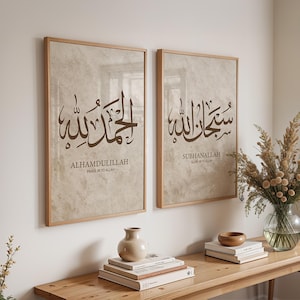 May include: Two framed art prints with Arabic calligraphy. The prints feature the phrases "ALHAMDULILLAH" and "SUBHANALLAH" in brown script on a beige background. The frames are a light wood color. The prints are displayed above a wooden console table.