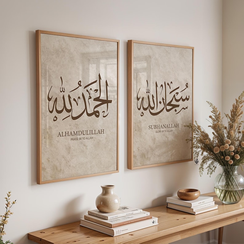 May include: Two framed art prints with Arabic calligraphy. The prints feature the phrases "ALHAMDULILLAH" and "SUBHANALLAH" in brown script on a beige background. The frames are a light wood color. The prints are displayed above a wooden console table.