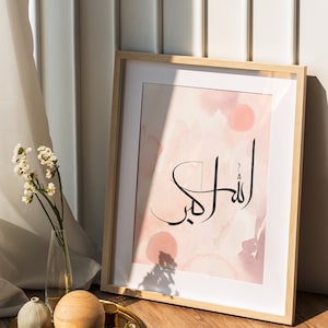 May include: A framed print with a pink watercolor background and black calligraphy that reads "Allah" in Arabic. The print is on a wooden floor with a gold tray, a small white vase, and a wooden ball.