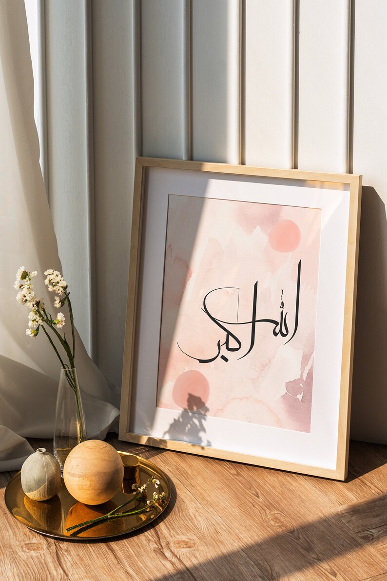 May include: A framed print with a pink watercolor background and black calligraphy that reads "Allah" in Arabic. The print is on a wooden floor with a gold tray, a small white vase, and a wooden ball.