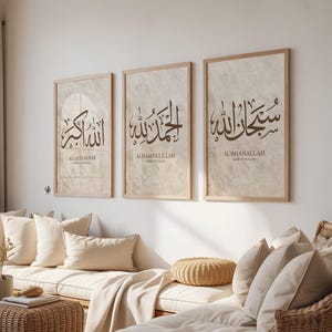 May include: Three framed art prints with Arabic calligraphy in brown script on a beige background. The prints are in light wood frames and are hung on a white wall. The text includes "ALLAHUAKBAR", "ALHAMDULILLAH", and "SUBHANALLAH".