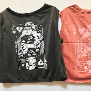 Puede incluir: Dos camisetas cortas sin mangas, una gris carbón y otra coral. Cada una presenta un gráfico blanco de una carta de reina con el texto "LET THEM EAT SKATE" y "QUEEN OF THE TRACK".