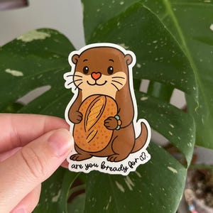 May include: A sticker featuring a cartoon otter holding a loaf of bread. The otter is brown with a pink heart-shaped nose and a blue bracelet. The text "are you bready for it?" is written below the otter.