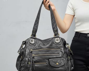 May include: Black leather handbag with multiple zippered compartments and a textured finish. The bag has two top handles and a shoulder strap. The hardware is a brass color. The bag is held up by a person.