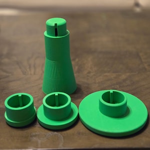 May include: A set of four bright green 3D-printed components. The set includes a tall, conical piece with a slotted top, and three circular pieces with raised, cylindrical centers. The pieces are arranged on a brown surface.