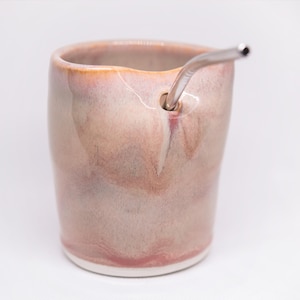 May include: A handmade ceramic mug with a unique, swirled glaze in shades of pink and peach. A stainless steel straw is inserted into the side of the mug. The mug has a rounded shape and a white base, perfect for enjoying cold beverages.