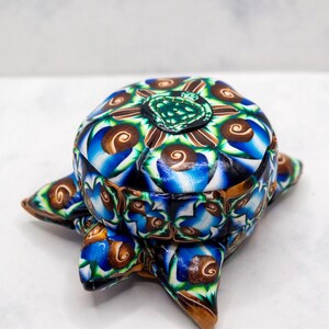 May include: A decorative box with a turtle shape, featuring a colourful pattern of blue, brown, green, and white swirls. The box has a lid and four feet, and is made of a glossy material. The box is a unique, handcrafted item.