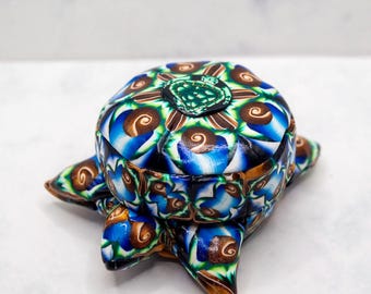 May include: A decorative box with a turtle shape, featuring a colourful pattern of blue, brown, green, and white swirls. The box has a lid and four feet, and is made of a glossy material. The box is a unique, handcrafted item.