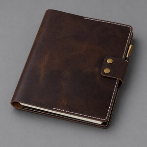 May include: A dark brown notebook cover with a smooth, matte finish and visible white stitching along the edges. The cover features a flap closure secured by two brass-colored metal snaps and includes a pen holder loop on the right side.