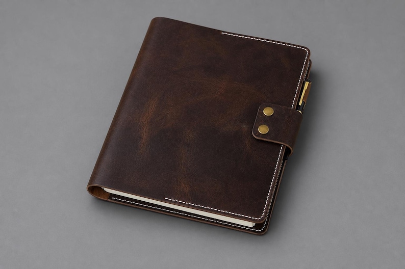 May include: A dark brown notebook cover with a smooth, matte finish and visible white stitching along the edges. The cover features a flap closure secured by two brass-colored metal snaps and includes a pen holder loop on the right side.
