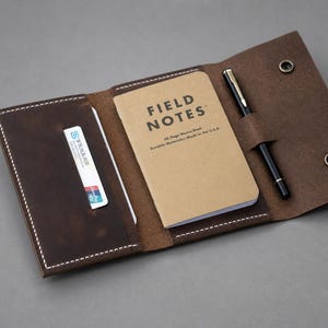 May include: A dark brown leather notebook cover with white stitching, featuring a card slot containing a white card, a tan Field Notes memo book, and a black pen held in a loop. The cover includes two metal snap buttons for secure closure.